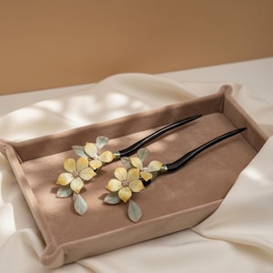 Puede incluir: Dos horquillas decorativas para el cabello con flores amarillas y detalles de hojas verdes. Las flores tienen un centro de perlas y están unidas a un palillo negro. Los accesorios para el cabello se exhiben en una bandeja beige.
