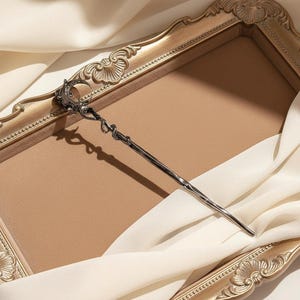 Op de afbeelding: Een zilverkleurige haarspeld met een decoratief maan- en slangenmotief. Het haaraccessoire is geplaatst tegen een goudkleurige achtergrond met een neutraal gekleurd interieur en witte stof.