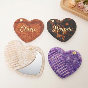 May include: Heart-shaped compact mirrors and combs in various colors and patterns. Two mirrors have names in gold script: "Claire" and "Harper." One mirror is open, revealing a mirror and comb. The other is closed. The combs are purple and cream.