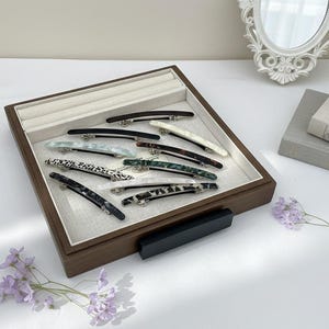 May include: A selection of barrette hair clips in a range of colours and patterns, including black, white, and tortoiseshell designs. The clips are displayed in a wooden tray with a light-coloured interior.