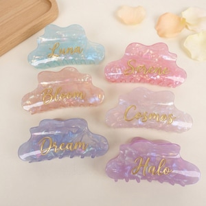May include: Six iridescent cloud-shaped hair claw clips in pastel colors: blue, pink, peach, lavender, and purple. Each clip has a gold script word: Luna, Serene, Bloom, Cosmos, Dream, and Halo. The clips are on a light background.