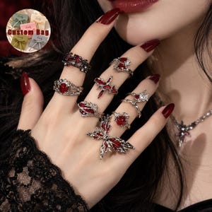Könnte beinhalten: Ein Set aus silberfarbenen Ringen mit roter Emaille und klaren Kristallakzenten in verschiedenen Formen, darunter ein großer Schmetterling, Herzen und abstrakte Designs. Die Ringe werden an den Fingern getragen, eine Kette ist sichtbar.