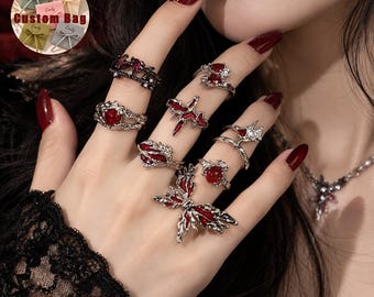 Peut inclure: Un ensemble de bagues argentées ornées d'émail rouge et de cristaux transparents aux formes variées, incluant un grand papillon, des cœurs et des motifs abstraits. Les bagues sont portées aux doigts et un collier assorti est visible.