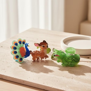 May include: Three decorative hair clips in the shape of animals. A peacock with blue, orange, and green feathers, a brown deer with white spots, and a green alligator. The clips are made of a translucent material.