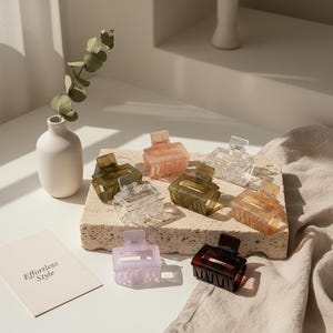 Puede incluir: Una colección de pequeñas pinzas para el cabello de colores en varios tonos, incluyendo transparente, rosa y marrón. Las pinzas están dispuestas sobre una superficie cuadrada texturizada. También hay un pequeño jarrón con una ramita de vegetación y una tarjeta con el texto "Effortless Style".