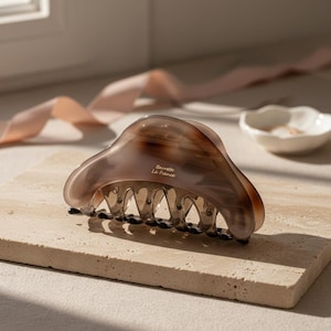 Puede incluir: Una pinza para el pelo marrón y beige con efecto veteado. La pinza tiene un diseño curvo con una serie de dientes y el texto "Barrette La France". La pinza está sobre una superficie de piedra beige.