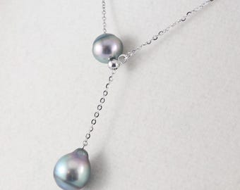 May include: A silver necklace featuring two iridescent gray pearls. The necklace has a delicate chain with a small pearl accent. The pearls are teardrop-shaped and round, with a lustrous finish. The necklace is displayed on a white surface.