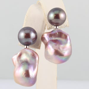 May include: A pair of pearl earrings. Each earring features a dark gray, round pearl stud and a larger, irregularly shaped, iridescent pearl in shades of pink, purple, and white. The earrings are displayed on a white stand.