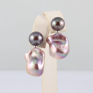 May include: A pair of earrings featuring a dark gray, round pearl stud and a large, iridescent, baroque pearl drop. The drop pearls have a mix of pink, purple, and gold hues. The earrings are displayed on a white stand.