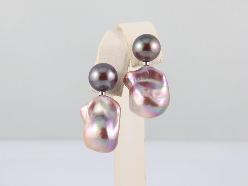 May include: A pair of earrings featuring a dark gray, round pearl stud and a large, iridescent, baroque pearl drop. The drop pearls have a mix of pink, purple, and gold hues. The earrings are displayed on a white stand.