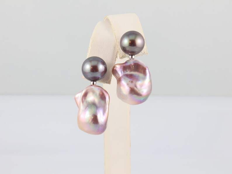 May include: A pair of pearl earrings. Each earring features a dark gray, round pearl stud, connected to a large, iridescent, baroque pearl. The baroque pearls have a pink and lavender hue, with a unique, irregular shape.