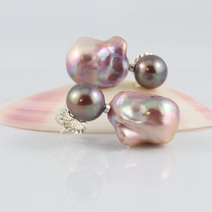 May include: A pair of pearl earrings with a unique design. Each earring features a large, iridescent, baroque pearl in shades of pink, purple, and gold, paired with a smaller, dark gray pearl. The earrings are set on silver posts.