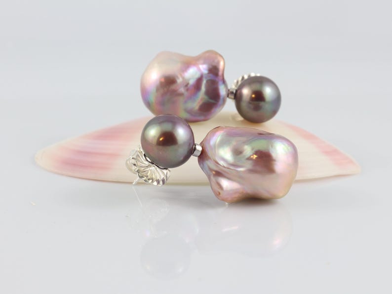 May include: A pair of pearl earrings with a unique design. Each earring features a large, iridescent, baroque pearl in shades of pink, purple, and gold, paired with a smaller, dark gray pearl. The earrings are set on silver posts.