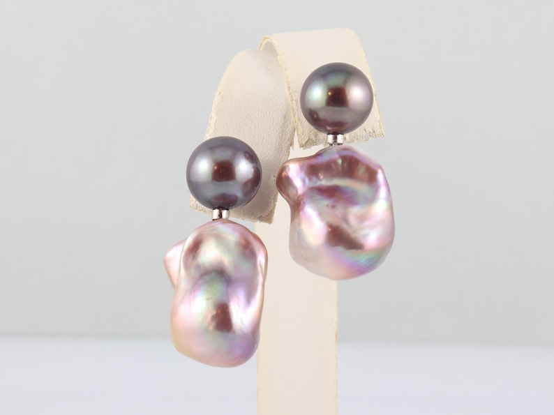May include: A pair of pearl earrings. Each earring features a dark gray, round pearl stud and a larger, irregularly shaped, iridescent pearl in shades of pink, purple, and white. The earrings are displayed on a white stand.