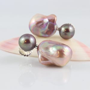 May include: A pair of pearl earrings with a dark gray, round pearl stud and a large, irregularly shaped, iridescent pearl drop. The earrings are displayed on a seashell with pink and white hues, set against a white background.