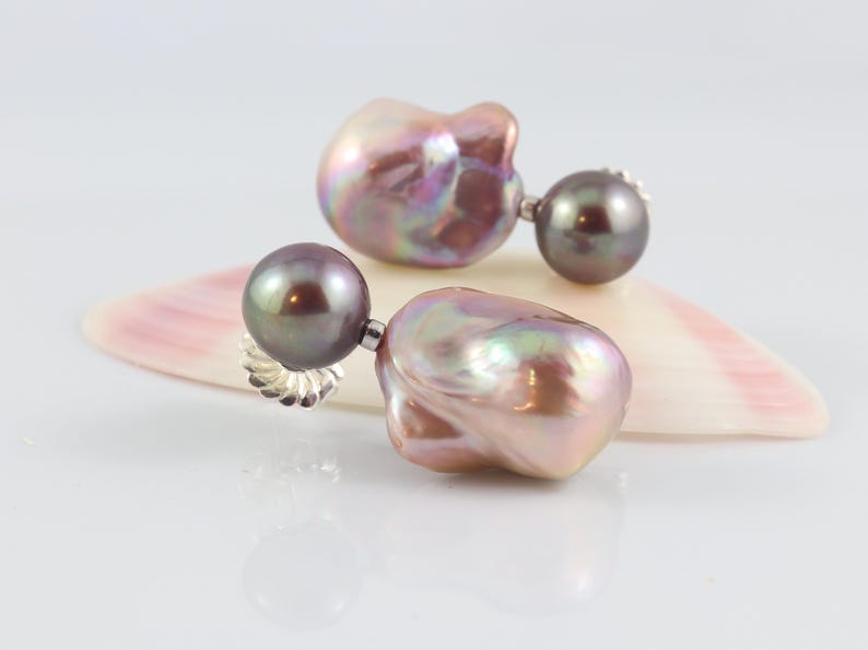 May include: A pair of pearl earrings with a dark gray, round pearl stud and a large, irregularly shaped, iridescent pearl drop. The earrings are displayed on a seashell with pink and white hues, set against a white background.