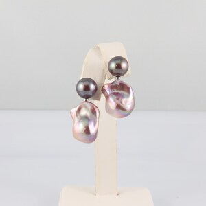 May include: A pair of pearl earrings. Each earring features a dark gray, round pearl stud and a larger, irregularly shaped, iridescent pearl in shades of pink, lavender, and silver. The earrings are displayed on a white stand.