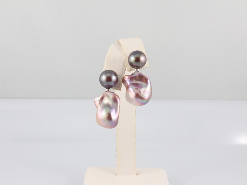 May include: A pair of pearl earrings. Each earring features a dark gray, round pearl stud and a larger, irregularly shaped, iridescent pearl in shades of pink, lavender, and silver. The earrings are displayed on a white stand.