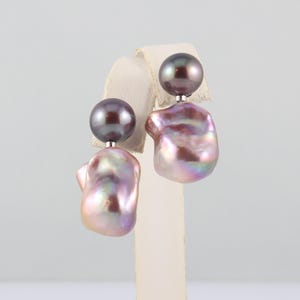 May include: A pair of pearl earrings. Each earring features a dark gray, round pearl stud and a large, irregular-shaped, iridescent pearl in shades of pink, purple, and white. The earrings are displayed on a white stand.