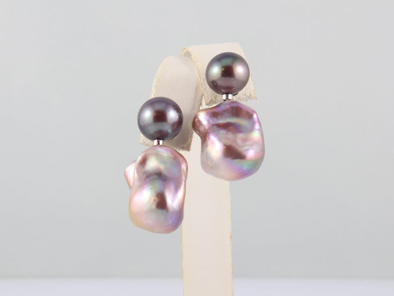 May include: A pair of pearl earrings. Each earring features a dark gray, round pearl stud and a large, irregular-shaped, iridescent pearl in shades of pink, purple, and white. The earrings are displayed on a white stand.