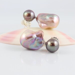 May include: A pair of pearl earrings with a dark gray round pearl and a large, iridescent, baroque pearl. The earrings are set on a white surface with a pink and white seashell.