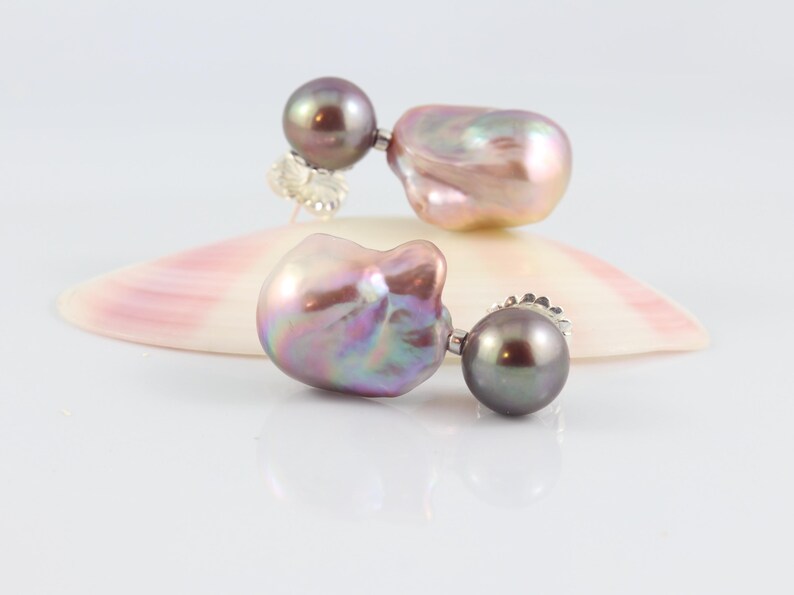 May include: A pair of pearl earrings with a dark gray round pearl and a large, iridescent, baroque pearl. The earrings are set on a white surface with a pink and white seashell.