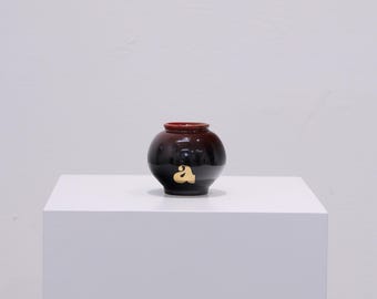 May include: A small, round ceramic vase with a dark brown base that fades to a reddish-brown rim. A gold-colored letter "a" is on the front. The vase is displayed on a white surface against a white background.