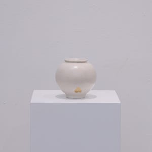 Puede incluir: Un pequeño jarrón redondo de cerámica blanquecina con un adorno dorado cerca de la base. El jarrón tiene una abertura ancha y está sobre un pedestal blanco. El fondo es una pared blanca lisa.