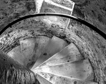 May include: A black and white image of a spiral staircase inside a stone tower. The wooden steps curve around a central point, with a dark metal handrail. The stone walls are rough and textured, and a small window is visible at the top.