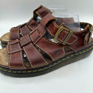 May include: Brown leather sandals with multiple straps and a buckle closure. The sandals have white stitching and a thick sole. The brand name "Dr. Martens" is visible on the buckle. The sandals are a size 9.