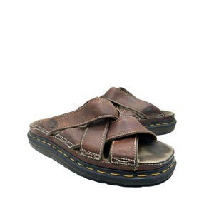 Può includere: Sandali in pelle marrone con cinturini incrociati e suola nera spessa. I sandali hanno cuciture bianche e una linea gialla attorno alla suola. Comodi e resistenti.