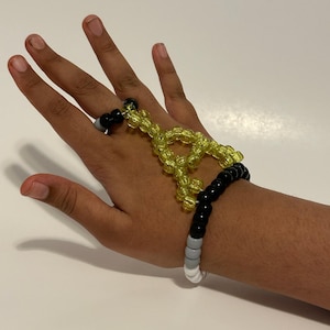 Könnte beinhalten: Ein Armband- und Ring-Set aus Perlen. Das Armband hat schwarze, graue und weiße Perlen. Der Ring hat schwarze und graue Perlen. Ein gelbes Perlendesign verbindet den Ring mit dem Armband. Der Schmuck befindet sich auf hellem Hintergrund.