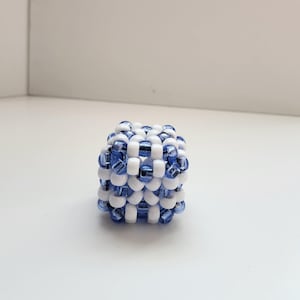Puede incluir: Un objeto en forma de cubo hecho de pequeñas cuentas redondas. Las cuentas son una mezcla de blanco y azul translúcido, dispuestas en un patrón de rejilla. El objeto está sobre una superficie blanca.