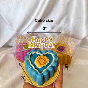 May include: A blue heart-shaped cake with a yellow rose on top, presented on a gold-colored plate. The cake is 3 inches wide and 3 inches tall, with a white border. A "Happy Birthday" topper is visible.