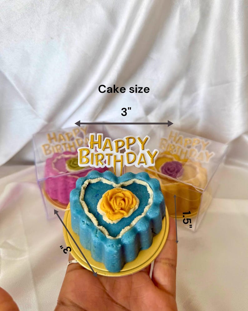 May include: A blue heart-shaped cake with a yellow rose on top, presented on a gold-colored plate. The cake is 3 inches wide and 3 inches tall, with a white border. A "Happy Birthday" topper is visible.