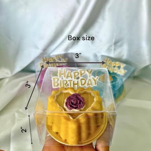 May include: A yellow cake with a purple rose decoration, inside a clear plastic box. The box is 3 inches by 3 inches by 2 inches. The words "HAPPY BIRTHDAY" are on top of the box.