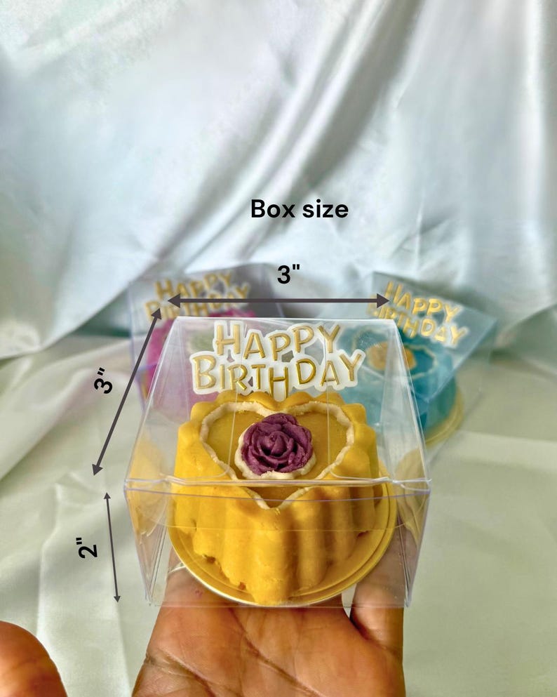May include: A yellow cake with a purple rose decoration, inside a clear plastic box. The box is 3 inches by 3 inches by 2 inches. The words "HAPPY BIRTHDAY" are on top of the box.