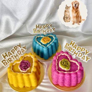 May include: Three heart-shaped cakes in yellow, blue, and pink, each with a flower and a "Happy Birthday" topper. The cakes sit on gold plates. A cat and dog image is in the background.
