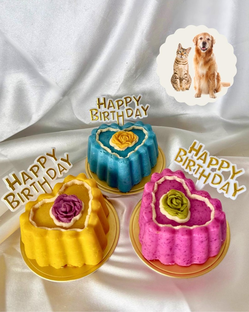 May include: Three heart-shaped cakes in yellow, blue, and pink, each with a flower and a "Happy Birthday" topper. The cakes sit on gold plates. A cat and dog image is in the background.