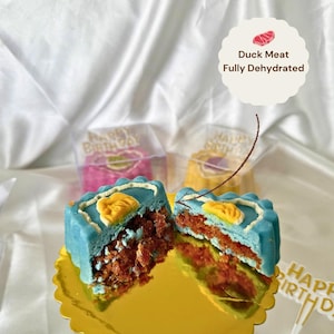 May include: A halved, blue-frosted cake with a yellow design and a red filling sits on a gold, scalloped plate. The cake is labeled "Duck Meat Fully Dehydrated". Two other cakes are visible in clear boxes, with a "Happy Birthday" cake topper.