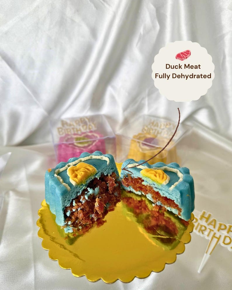 May include: A halved, blue-frosted cake with a yellow design and a red filling sits on a gold, scalloped plate. The cake is labeled "Duck Meat Fully Dehydrated". Two other cakes are visible in clear boxes, with a "Happy Birthday" cake topper.