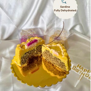 May include: A yellow, round, partially sliced cake with a decorative edge, revealing a layered interior. The cake sits on a gold scalloped plate. The text "Sardine Fully Dehydrated" is in the image. A "Happy Birthday" cake topper is also visible.