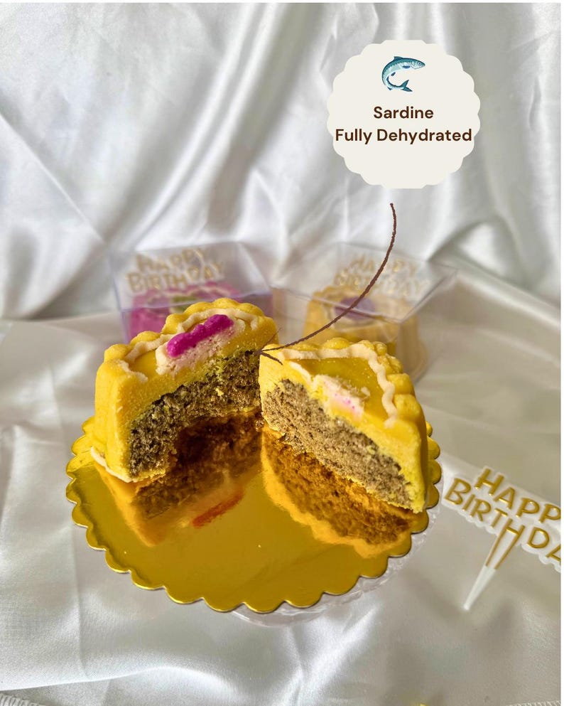 May include: A yellow, round, partially sliced cake with a decorative edge, revealing a layered interior. The cake sits on a gold scalloped plate. The text "Sardine Fully Dehydrated" is in the image. A "Happy Birthday" cake topper is also visible.