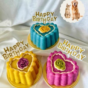 May include: Three heart-shaped cakes in blue, yellow, and pink, each with a different colored rose and a gold "Happy Birthday" topper. The cakes sit on gold plates. A small image of a cat and a dog is in the background.