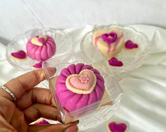 May include: A close-up of a pink and white heart-shaped dessert in a clear box. The dessert has a heart-shaped design on top. The text "With Real Meat!" is visible in the background. Other desserts are on glass plates.