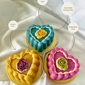 May include: Three heart-shaped cakes in yellow, blue, and pink, each with a flower design in the center. The cakes sit on small, round, yellow plates. The text labels indicate the colors are turmeric, spirulina, and dragonfruit.