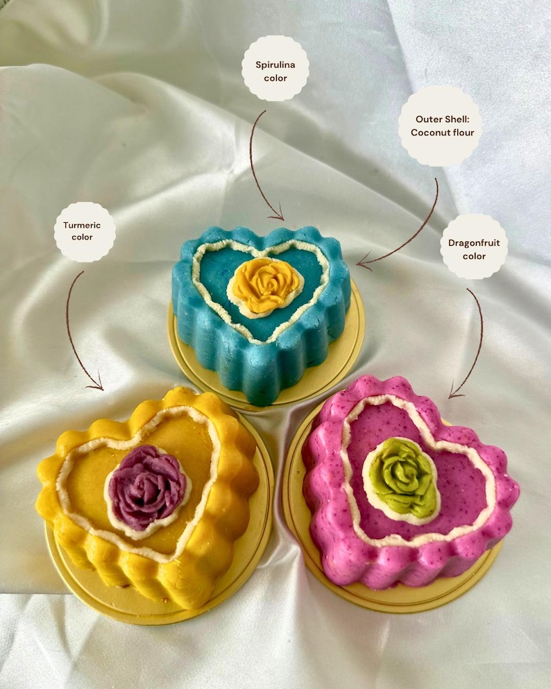 May include: Three heart-shaped cakes in yellow, blue, and pink, each with a flower design in the center. The cakes sit on small, round, yellow plates. The text labels indicate the colors are turmeric, spirulina, and dragonfruit.