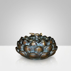 May include: A bronze-colored, lotus-shaped decorative object. The object has a detailed, layered petal design and a small, bird-like finial on top. The surface has a metallic sheen, and the overall form is symmetrical and elegant.