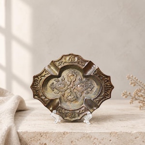 May include: An oval-shaped metal tray with a dark, weathered bronze finish. The surface features an intricate, embossed floral pattern in the center and along the scalloped edges, with four distinct indentations positioned around the perimeter.