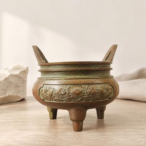 May include: A bronze-toned metal incense burner featuring a rounded body with three short legs and two upright handles. The exterior surface displays a horizontal band with embossed floral motifs and a weathered, greenish patina finish.